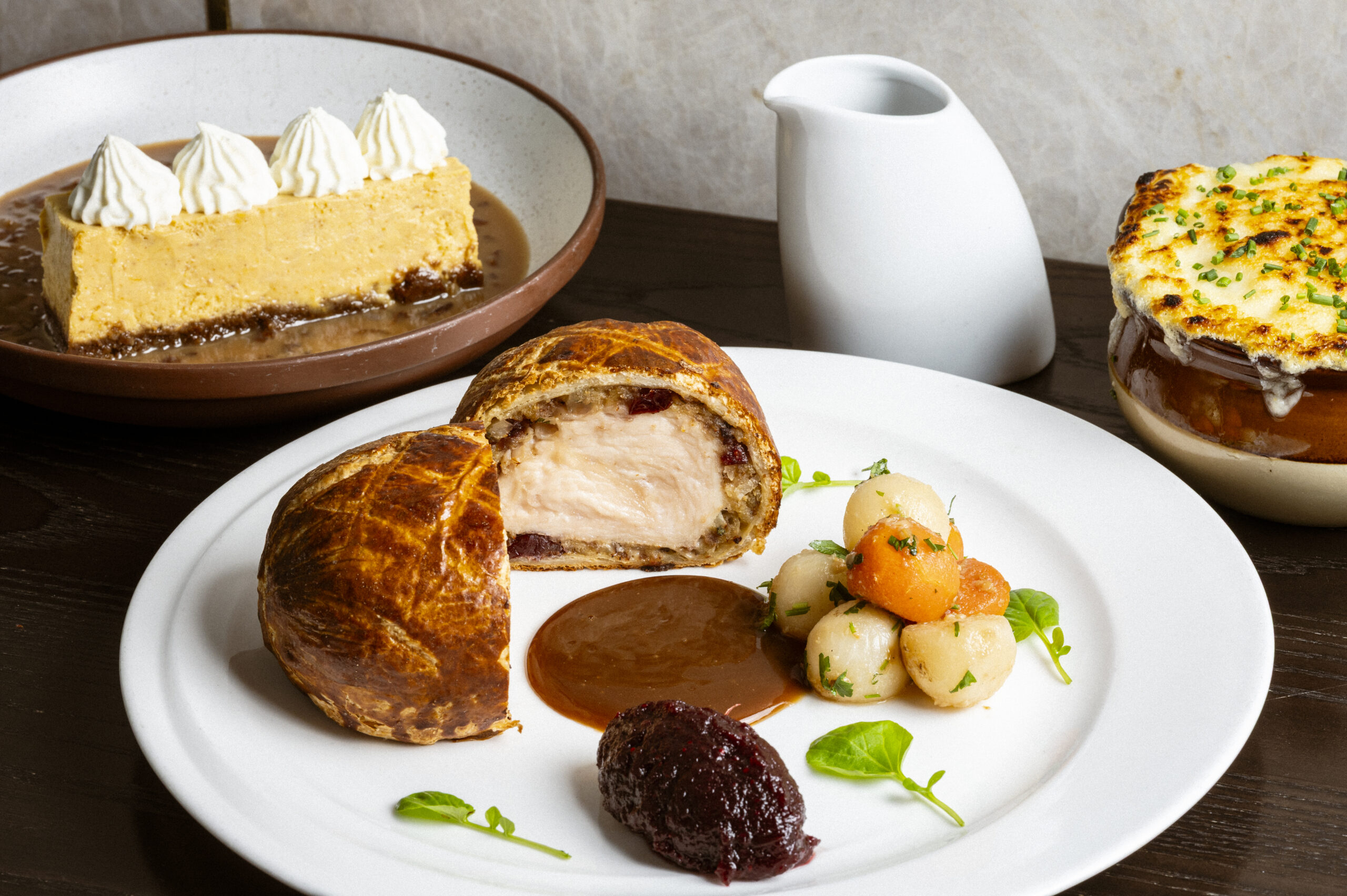 A plate featuring a sliced meat pasty, roasted vegetables, and sauce, with a dessert and soup bowl in the background.
