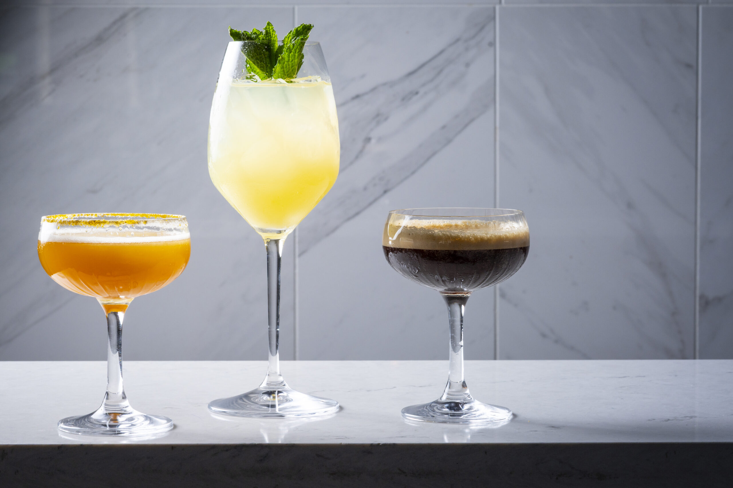 Three different cocktails in stemmed glasses lined up on a marble countertop.